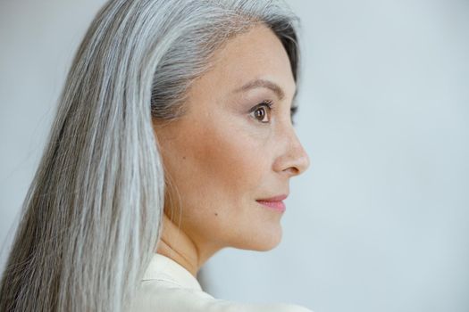 Middle aged Asian woman with beautiful straight hoary hair poses on light grey background in studio closeup side view. Mature beauty lifestyle