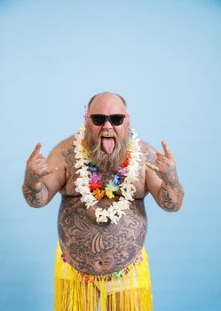 Funny bearded man with overweight in decorative grass skirt and flowers garland shows Horns gestures and tongue on light blue background in studio