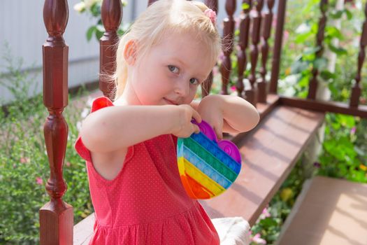 Cute caucasian fair hair girl in pink dress is playing and showing funny trendy silicone antistress colorful toy popit. Push Popper fidget toy is the ideal toy to get rid of your frustration or stress.