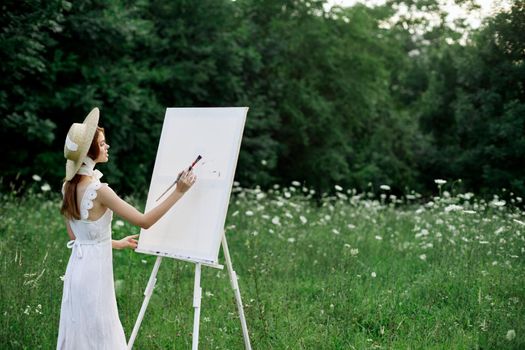 A woman in a white dress in a field with flowers paints a picture. High quality photo