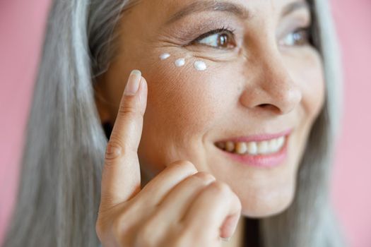 Beautiful grey haired Asian lady applies antiaging cream under eye on pink background in studio closeup. Mature beauty lifestyle