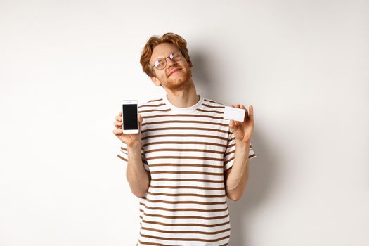 Shopping and finance concept. Pleased young man with red hair smiling from satisfaction, showing smartphone blank screen and credit card, white background.