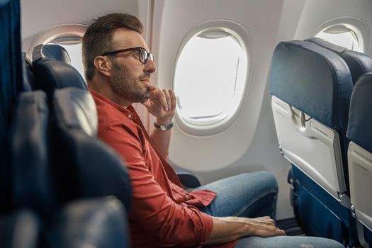 Handsome caucasian man in casual wear and glasses looking away, sitting on the plane near the window. Relax, travel, vacation, transportation concept