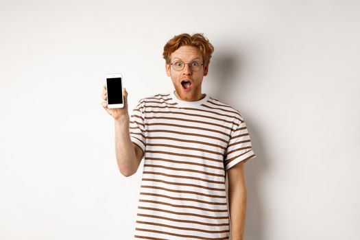 Technology and e-commerce concept. Surprised and shocked redhead guy checking out online promotion, showing blank smartphone screen and drop jaw, white background.