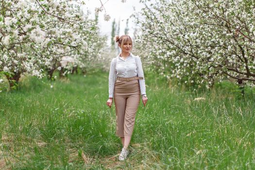 Beautiful young blonde woman in white shirt with backpack posing under apple tree in blossom and green grass in Spring garden