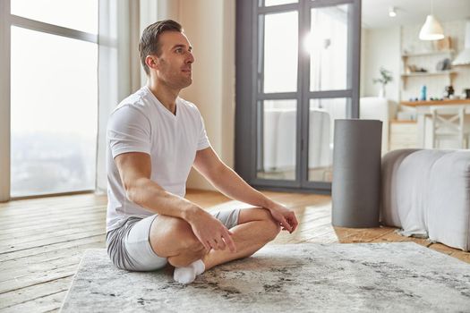 Happy young man is sitting in lotus position in living room and relaxing after training on mat indoors