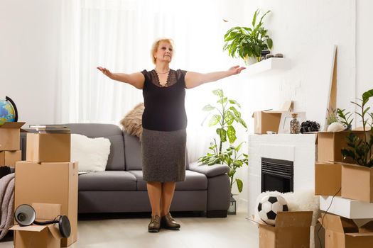 Happy excited mature retired middle aged woman standing in modern living room interior alone with arms outstretched enjoy freedom and wellbeing feel motivated at new furnished renovated home concept