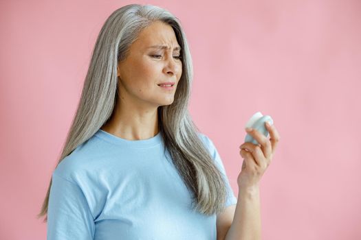 Doubting hoary haired Asian woman looks at sonic facial cleansing brush on pink background in studio. Mature beauty lifestyle