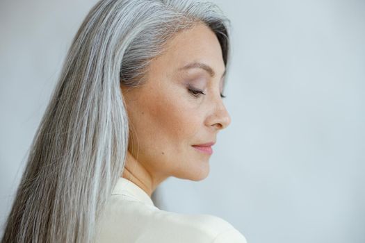 Pretty middle aged Asian woman with loose straight grey hair poses on light background in studio closeup side view. Mature beauty lifestyle