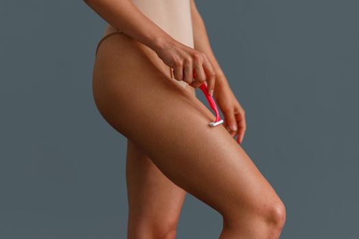 Close up shot of woman shaving her leg using razor, standing over gray studio background. Beauty, body care concept