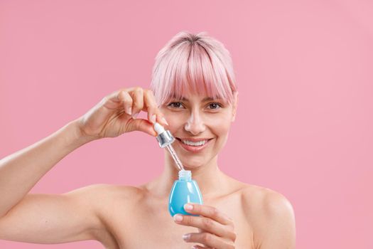 Portrait of smiling young woman with pink hair and bare shoulders looking at camera, holding bottle of serum, posing isolated over pink background. Beauty, cosmetics and skincare concept