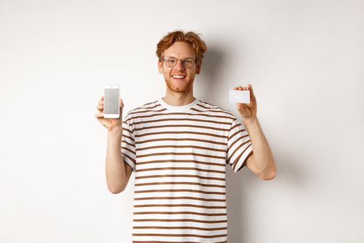 Shopping and finance concept. Young man showing blank mobile screen and plastic credit card, smiling at camera, white background.