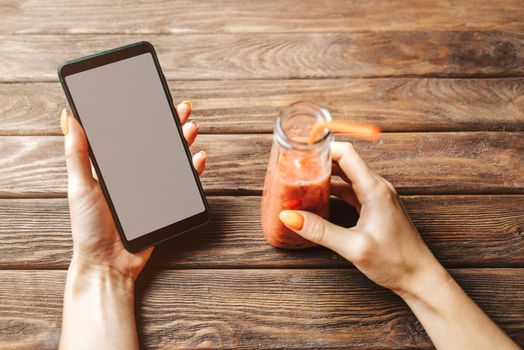 Woman holding smartphone with blank screen and bottle of fresh smoothie drink at wooden table. Point of view in first person.