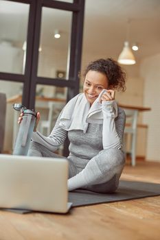 Low angle of jolly slim woman sitting on mat with towel and bottle of water before laptop