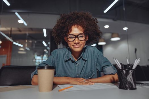 Smiling attentive woman wearing glasses and sitting at workplace with cup of coffee and document at the table