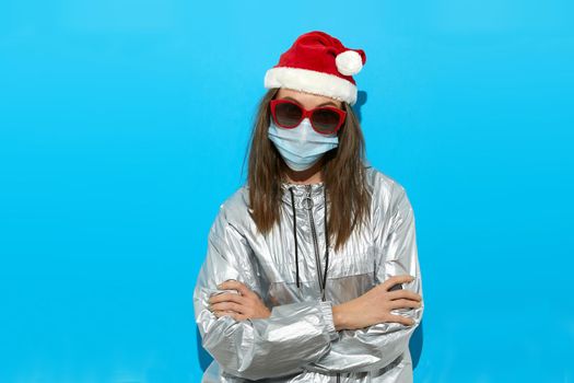 Serious female wearing Santa hat and protective mask and red sunglasses standing on blue background in studio and looking at camera Arms crossed
