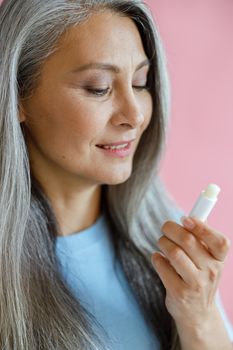 Smiling grey haired Asian lady in blue t-shirt looks at lip balm on pink background in studio. Mature beauty lifestyle