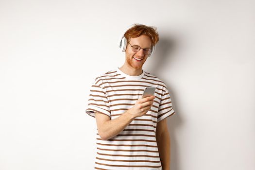 Technology concept. Young redhead hipster in glasses, listening music in headphoes, reading message on smartphone, white background.