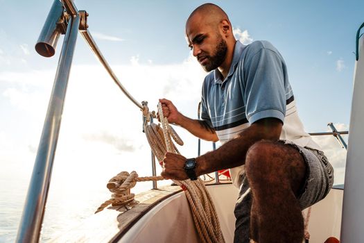 Young african american sailor tying ropes on sailboat in the sea on sunset, close up