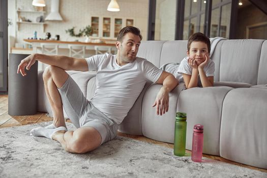 Cheerful kid is lying on sofa while dad is sitting on floor with sport bottles of water during active day at home