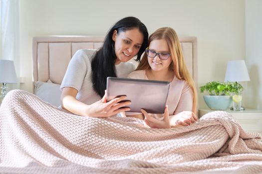 Happy mom and preteen daughter hugging together looking at screen of digital tablet, sitting at home in bed under blanket. Family, children, relationships, home, lifestyle, people concept