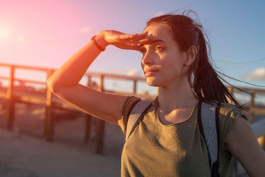 Fashionable slender young brunette girl with backpack posing on a background wooden walkways and sand beach at sunset.