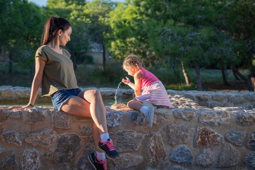 Beautiful mother and her little daughter outdoors. Beauty Mum and her Child playing with water in Park together at sunset. Outdoor Portrait of happy family. Mother's Day.