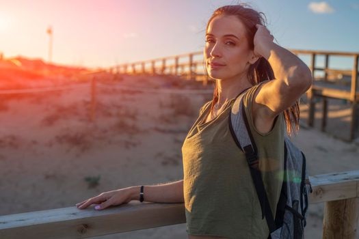 Fashionable slender young brunette girl with backpack posing on a background wooden walkways and sand beach at sunset.