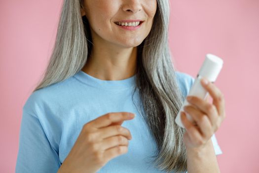 Smiling grey haired Asian lady holds bottle of cosmetic product on pink background in studio closeup. Mature beauty lifestyle