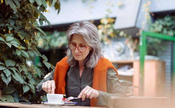 Grey haired senior woman eats delicious dessert near cup of drink sitting at table on outdoors cafe terrace on autumn day