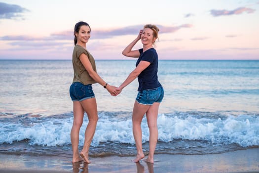 leisure and friendship concept - two happy smiling young women or best friends at seaside holding hands at sunset standing in water.