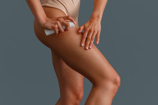 Close up shot of woman applying foam or soothing body lotion on her leg after depilation, standing over gray studio background. Beauty, body care concept