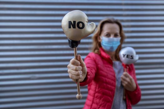 Young woman in protective sterile medical mask on her face, on striped background, holding small boxing gloves with Yes No signs. America president election, pandemic coronavirus concept.