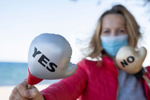 Young woman in protective sterile medical mask on sea background, holding small boxing glove with Yes No signs. America president election, pandemic coronavirus concept.