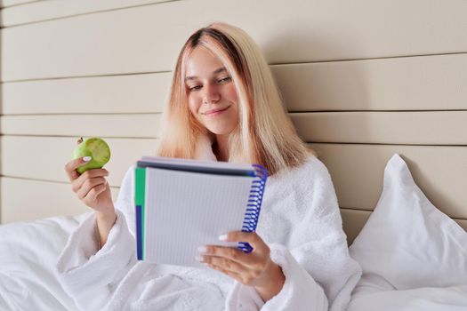 Teenager girl student with green apple and school notebook. Young female learns at home, sitting in bed eating natural fruits