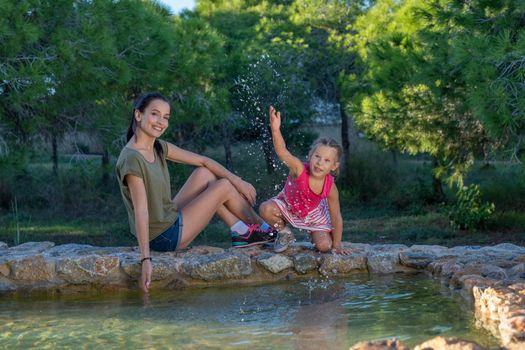 Beautiful mother and her little daughter outdoors. Beauty Mum and her Child playing with water in Park together at sunset. Outdoor Portrait of happy family. Mother's Day.