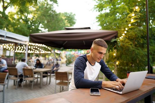 Young man sitting at table and typing on laptop keyboard while working in outdoor cafe, close up