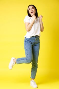 Full size shot of happy asian woman dancing and jumping from happiness, winning and celebrating victory, posing over yellow background.