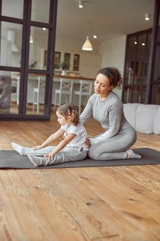 Calm mother is helping her little girl going stretching exercises on mat in living room