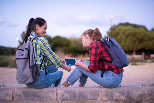 two young women students with backpack holding and looking at the tablet and smiling. Girl shows something on the screen.