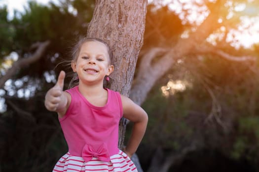 Closeup portrait of a cute attractive little child girl showing thumb up in the park with dark trees on background.