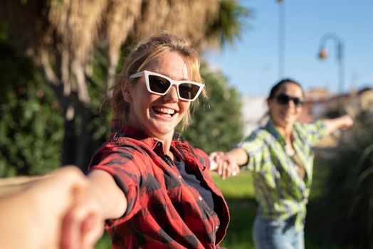 Pretty young caucasian woman in sunglasses pulling along by the hand invitingly with a happy smile as her friend on the background. Sunny summer day.