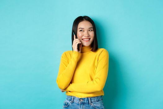 Attractive korean woman in yellow sweater, having conversation and smiling, talking on mobile phone, looking left at copy space, standing over blue background.