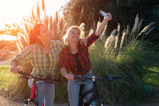 Two pretty young caucasian girls making selfie photo with smartphone. Best friends enjoying a day on bikes. Sunny summer evening.