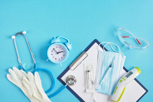 Alarm clock and medical equipment on a blue background place copy top view stethoscope non-contact thermometer face mask syringe safety glasses and gloves