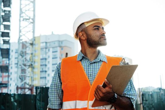 Young man engineer in workwear standing in construction site with clipboard, close up portrait