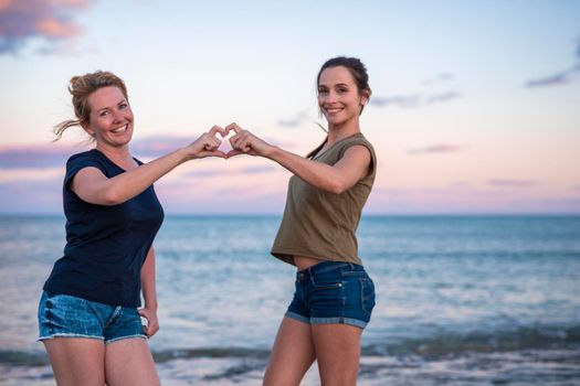 leisure and friendship concept - two happy smiling teenage girls or best friends at seaside making hand heart gesture. selective focus.