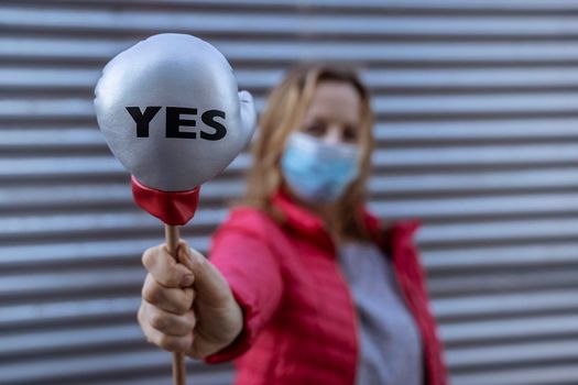 Young woman in protective sterile medical mask on her face, on striped background, holding small boxing glove with Yes sign. America president election, pandemic coronavirus concept.