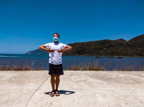 Coronavirus COVID 19. Young beautiful man doing sport performing workouts exercises in the park near sea during quarantine. Active life in surgical sterilizing face mask protection.