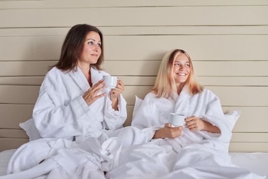 Happy mother and teenage daughter drinking coffee talking. Women in white bathrobes, resting sitting in bed, vacation together. Communication between parent and adolescent child, family, relationship